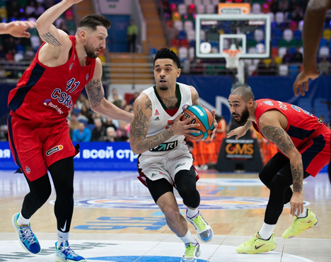 Игрок в белой форме ведет баскетбольный мяч, находясь между двумя соперниками в красной форме на площадке. Вокруг видны трибуны с болельщиками