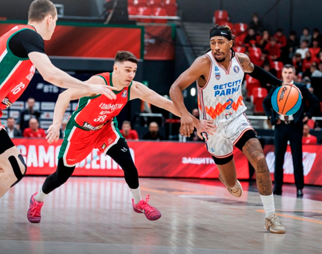 На изображении запечатлен момент баскетбольного матча: игрок в белой форме с мячом уходит от двух соперников в красной форме, пытающихся помешать. На заднем плане видны трибуны и рекламные баннеры